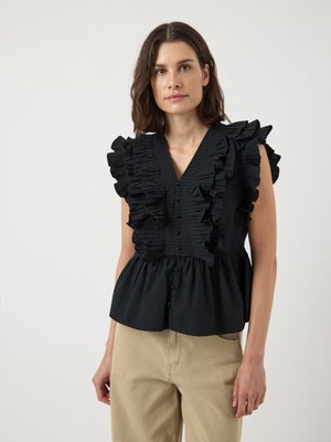 Woman wearing a sleeveless black blouse with ruffled pleats at the front and beige pants, standing against a plain background.