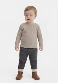 Batole stojí, má na sobě béžové tričko s dlouhým rukávem, tmavě šedé kalhoty s manžetami a hnědé kotníkové boty na jednobarevném bílém pozadí.