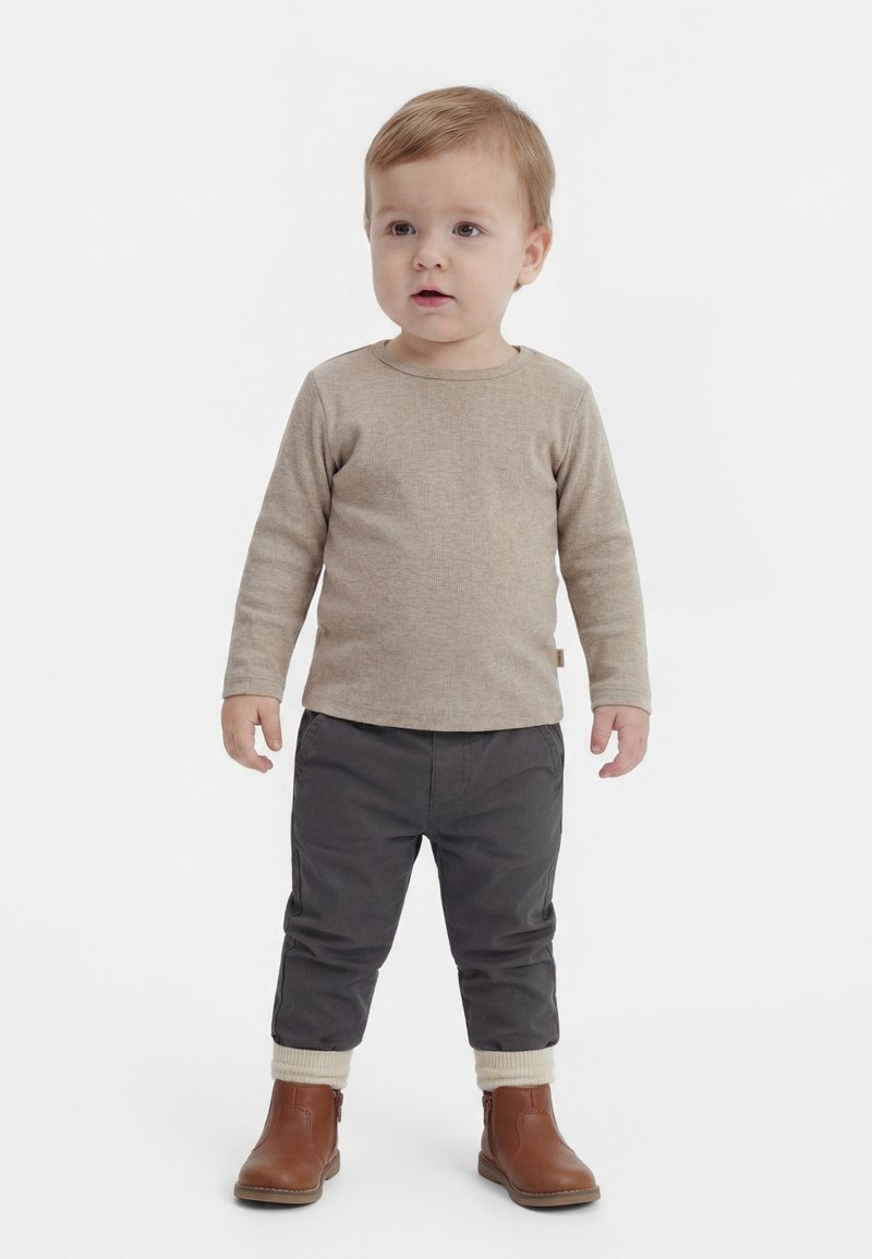 Batole stojí, má na sobě béžové tričko s dlouhým rukávem, tmavě šedé kalhoty s manžetami a hnědé kotníkové boty na jednobarevném bílém pozadí.