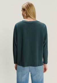 Kobieta z długimi blond włosami, nosząca złote kolczyki w kształcie kół, ciemnozielony, długi rękaw, oraz jasnoniebieskie dżinsy, stojąca tyłem na jednolitym tle.