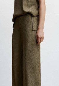 Tenue vert olive composée d'un haut texturé et froissé et de pantalons à jambes larges avec un motif diagonal subtil, portée avec une main sur le côté du pantalon.