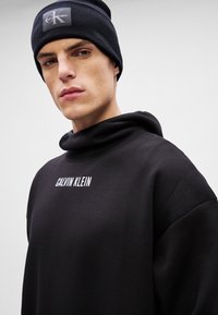 Čierna mikina s vysokým golierom, na ktorej je v bielom texte napísané "CALVIN KLEIN". V kombinácii s čiernou rebrovanou čiapkou so značkovou nášivkou.