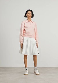 Sudadera rosa claro con cuello y botones, combinada con una falda blanca plisada y zapatillas blancas. Textura suave y diseño casual.