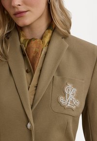 Blazer beige en tissu texturé, avec une poche sur la poitrine gauche et une broderie blanche complexe. Une écharpe à motifs marron et verte est visible.