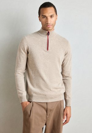 Beige quarter-zip sweater with red and blue accents, soft texture, long sleeves, ribbed cuffs, and a fitted waistband. Paired with brown trousers.