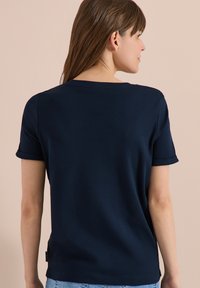 Mujer con pelo liso y castaño que lleva una camiseta de manga corta azul marino, vista desde atrás sobre un fondo beige.