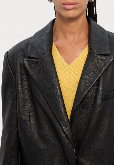 Black leather blazer with a textured finish, worn over a yellow ribbed V-neck sweater. The blazer features wide lapels and a single pocket.