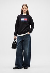 Musta huppari, jossa on sininen ja punainen "TOMMY JEANS" -logo, rento malli. Yhdistettynä leveälahkeisiin tummiin farkkuihin ja pieneen mustaan käsilaukkuun.