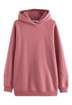 Femme en sweat-shirt rose et short bordeaux tenant un casque sans fil gris dans une pièce lumineuse et blanche avec de grandes fenêtres.