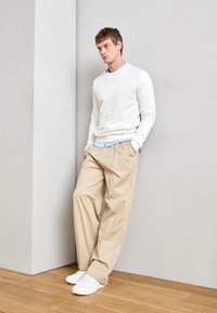 White crewneck sweatshirt over a light blue collared shirt, paired with beige loose-fit trousers and white sneakers, against a gray wall.