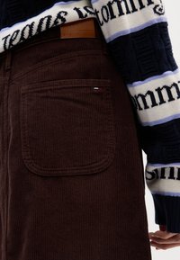 Pantaloni in velluto a coste marroni con una tasca posteriore che presenta un piccolo logo rosso, bianco e blu. Il capo superiore è un maglione a motivi in colori scuri.