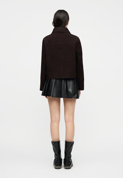 Γυναίκα που στέκεται πλάτη, φορώντας ένα σκούρο καφέ μπουφάν, μαύρη πλισέ δερμάτινη φούστα και μαύρες μπότες μέχρι τη μέση της γάμπας με λεπτομέρειες αλυσίδας.
