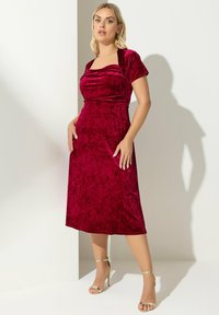 Vestido midi de terciopelo en rojo profundo, con mangas cortas, escote cuadrado y una textura sutil. Complementado con tacones dorados con tiras.