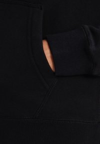 Sweat à capuche noir avec une poche avant kangourou, des poignets côtelés et une texture douce. Les surpiqûres visibles et le contraste des matériaux rehaussent le design.