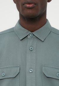 Nærbilde av en persons nedre del av ansiktet og nakke som har på seg en grønn skjorte med knapper, to brystlommer og krage.