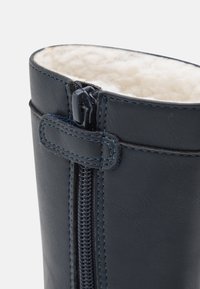 Botte en cuir bleu foncé avec coutures apparentes, une fermeture éclair latérale et une doublure douce de couleur crème en haut.