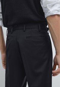 Tailored black trousers with a smooth, matte texture, featuring two back pockets and belt loops. Worn with a black knit vest over a white shirt.
