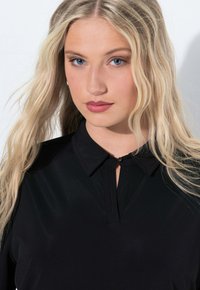 Blusa nera con colletto e piccolo gancio, texture morbida e dettagli di pieghe sottili nel collo. Capelli raccolti in onde morbide.