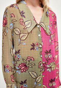 Blouse en dentelle florale avec un grand col, des boutons sur le devant et un design en blocs de couleurs avec des sections verte et rose.