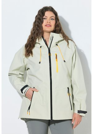 Kvinde med langt, bølget hår iført en lys beige jakke med sorte og gule lynlåsdetaljer, hænderne i lommerne, stående foran en hvid væg.