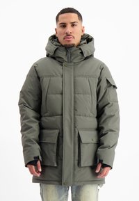 Man in een groene gewatteerde winterjas met rits en capuchon, met klepzakken en klittenband voor de polsen, staande tegen een effen witte achtergrond.