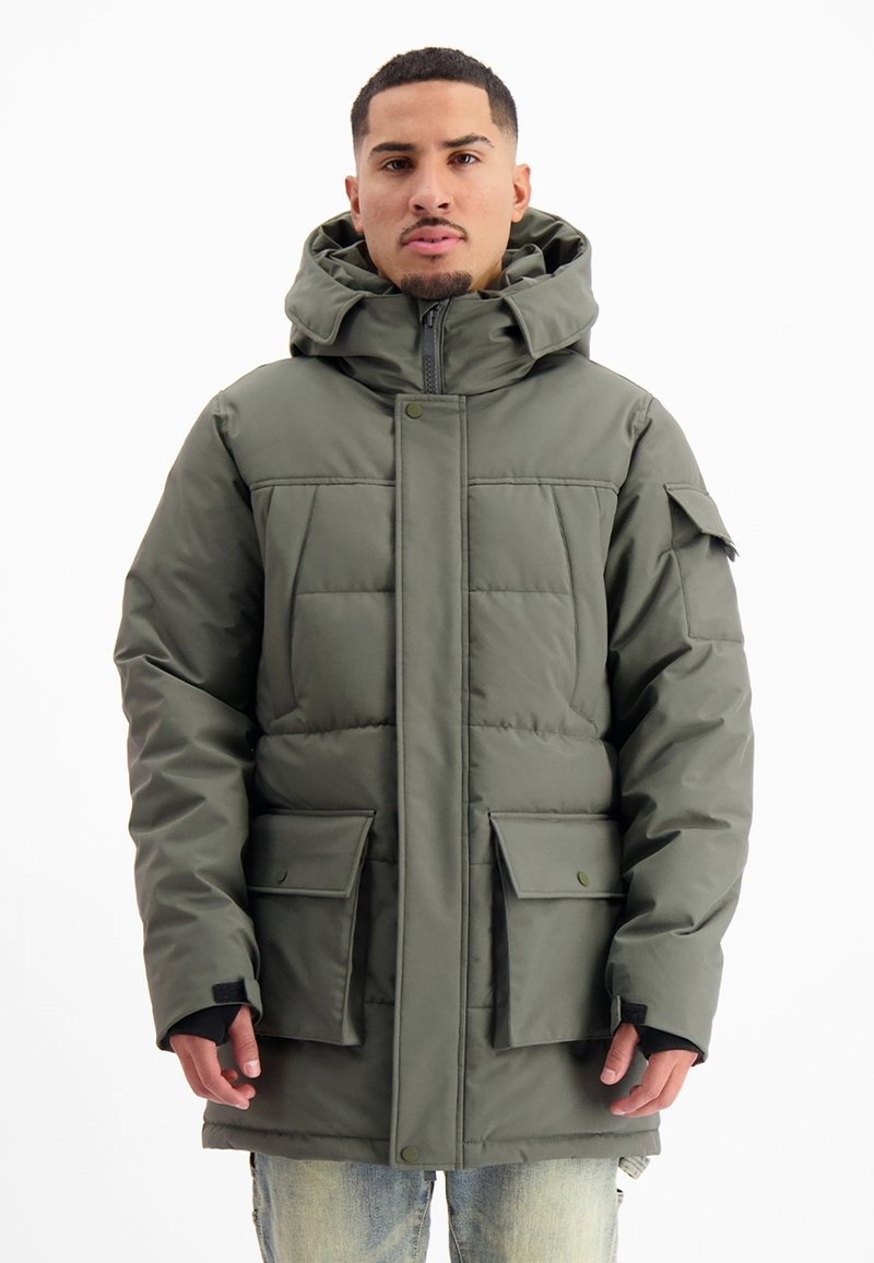 Man in een groene gewatteerde winterjas met rits en capuchon, met klepzakken en klittenband voor de polsen, staande tegen een effen witte achtergrond.