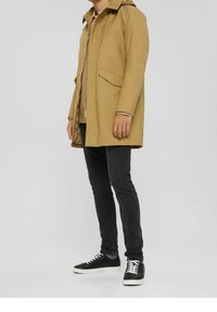 Wodoodporna parka w kolorze khaki z kapturem, zamkiem z przodu i dwoma bocznymi kieszeniami. Połączona z czarnymi dżinsami i czarnymi trampkami z białymi sznurowadłami.