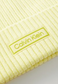 Yellow knitted beanie with vertical ribbing and a textured label showing "Calvin Klein" in light green on a rectangular background.