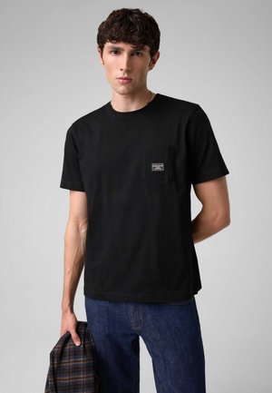 Junger Mann mit kurzen lockigen Haaren, der ein schwarzes T-Shirt und blaue Jeans trägt und eine zusammengefaltete karierte Jacke in der linken Hand hält.