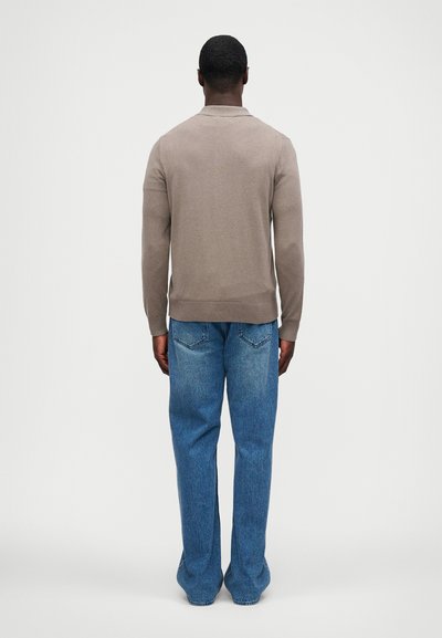 Μπεζ πουλόβερ με ριμπ τελείωμα, φορεμένο πάνω από μπλε τζιν σε χαλαρή εφαρμογή. Θέα από πίσω που αναδεικνύει τον απλό, καθαρό σχεδιασμό και την ομαλή υφή.