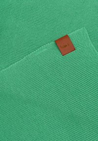 Teksturowana zielona dzianina z brązowym skórzanym naszywką z wytłoczonym logo. Materiał jest równomiernie tkany, prezentując spójną strukturę.