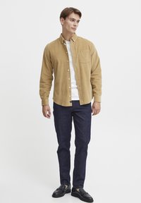 Lichtbruine corduroy overhemd met knopen en een enkele borstzak, gedragen over een wit T-shirt, gecombineerd met donkerblauwe spijkerbroeken en zwarte schoenen.