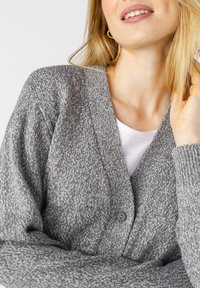Cardigan grigio a maglia caratterizzato da un design a scollo a V, chiusura a bottoni e tessuto testurizzato. Indossato sopra una camicia bianca.