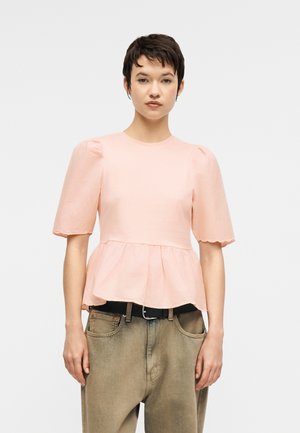 Donna con capelli corti scuri che indossa una camicetta rosa chiaro con maniche a sbuffo e orlo smerlato, abbinata a pantaloni beige a vita alta con cintura nera.