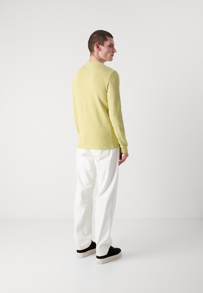 Lys gul ribbet langærmet top, hvide afslappede bukser og sorte sneakers med beige såler. Simpelt design, glat tekstur.