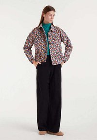 Chaqueta con estampado floral en colores vibrantes, superpuesta a un cuello de tortuga azul verdoso, combinada con pantalones de pierna ancha negros y calzado beige.