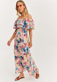 Kobieta w kwiecistej maxisukience na ramiączkach, brązowych sandałach i okularach przeciwsłonecznych w pomarańczowym odcieniu, stojąca na jednolitym tle.