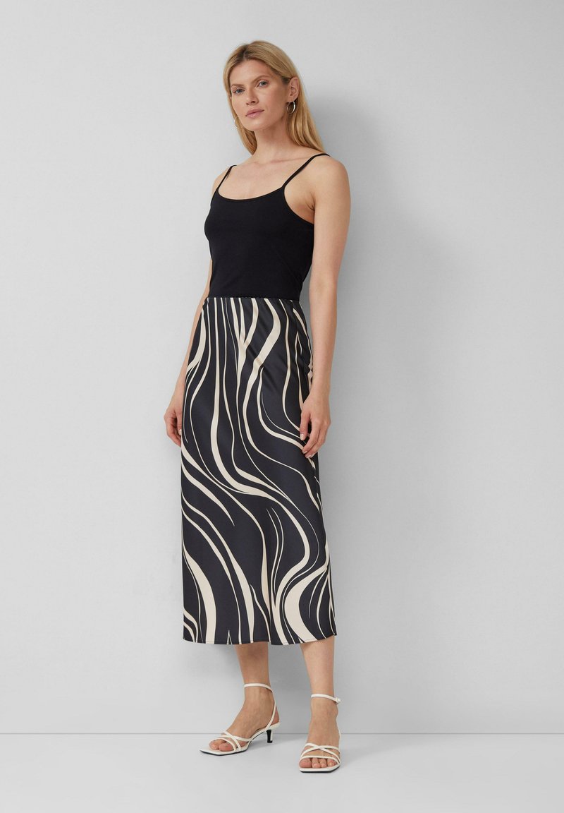 Zwarte getailleerde tanktop gecombineerd met een lange rok met een draaiend crèmekleurig patroon op een zwarte achtergrond. Witte sandaaltjes met bandjes maken de look compleet.