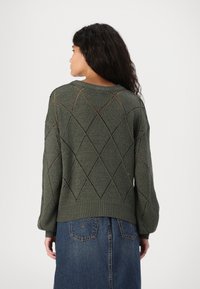 Kiefergrüner gestrickter Pullover mit Rautenmuster, Ballonärmeln und geripptem Saum. Getragen über einem Jeansrock.