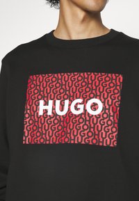 Sudadera negra con un gran diseño gráfico que presenta letras rojas y negras que dicen "HUGO" sobre un patrón repetido. Material de mezcla de algodón.