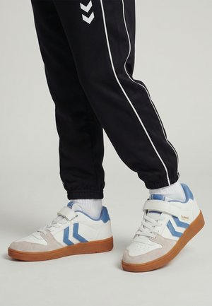 Person wearing black track pants and white sneakers with blue accents and gum soles, standing on a light gray floor.