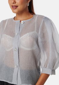 Blusa leggera grigia, trasparente, con una finitura testurizzata, scollo rotondo e grandi bottoni decorativi. Maniche leggermente a sbuffo aggiungono volume.