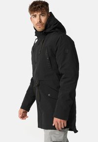 Parka noire avec capuche, dotée d'une fermeture à boutons, d'une taille à cordon de serrage et de deux poches frontales en biais. Texture de tissu lisse, design durable.