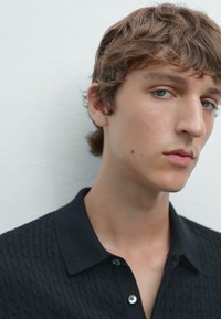 Polo nero con un motivo testurizzato, caratterizzato da un colletto e una chiusura a un bottone. Il tessuto sembra morbido e leggero.
