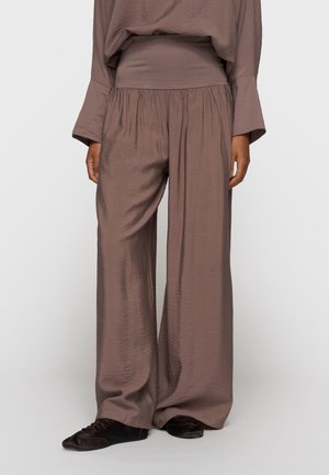 Brązowe spodnie o szerokich nogawkach z marszczonym elastycznym pasem, noszone z dopasowanym topem i czarnymi miękkimi płaskimi butami, przedstawione na stojącej osobie.