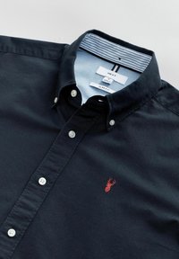 Camisa de algodón navy ajustada con botones blancos, que presenta un logotipo de cangrejo bordado en rojo en el lado izquierdo del pecho y un cuello interno a rayas.