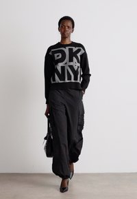 Melns džemperis ar pelēko "DKNY" uzrakstu, apvienots ar brīvi pieguļošām melnām kravu biksēm. Modelis tur melnu rokassomu un valkā šauras papēžkurpes.