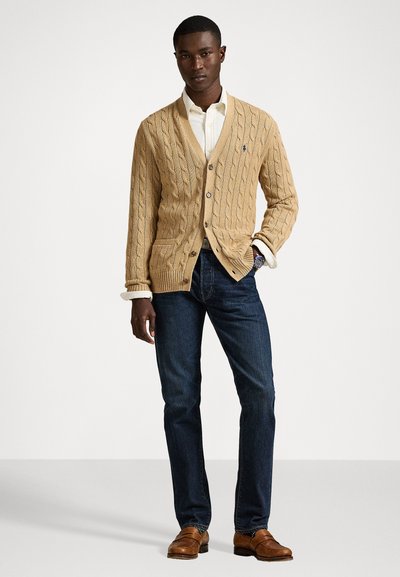 Bēšas krāsas adīts kardigans ar V veida kakla izgriezumu, pogu aizdari un divām priekšējām kabatām, kombinēts ar tumšiem džinsiem un brūnām kurpēm.