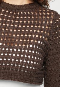 Pull en crochet marron avec un motif ajouré porté par-dessus un vêtement blanc, présentant une couture texturée et un col rond.