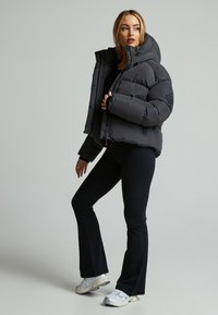 Giacca a vento nera con colletto alto, presenta un logo tonal, bottoni a scatto e un design cropped. Il modello indossa pantaloni neri e sneakers.
